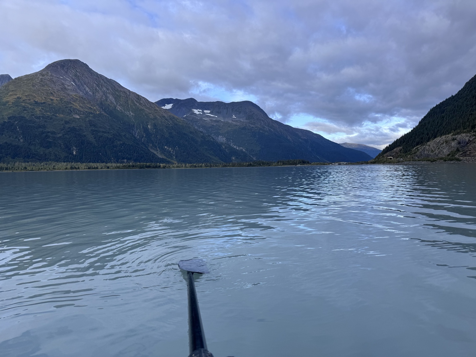 A level view across Portage Lake toward mountains beneath blue-gray clouds, with a rowing oar in the water